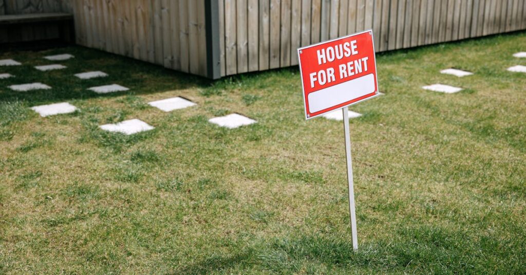 A red 'House for Rent' sign stands on a grassy lawn beside a wooden house exterior.